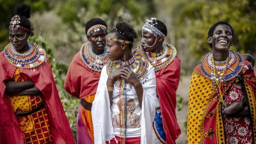Five members of the Masai community stand together wearing brightly colored traditional clothing and laughing.